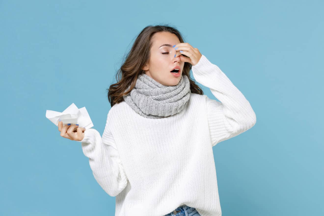 Giovane donna influenzata durante la stagione fredda, con maglione bianco e sciarpa mentre tiene in mano un fazzoletto.